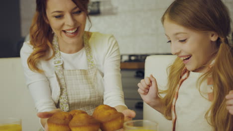 Young-pretty-mother-in-the-apron-putting-a-plate-with-muffins-on-the-kitchen-table-in-front-of-her-happy-daughter.-Indoors